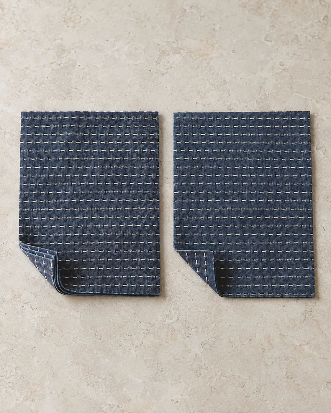 Two indigo cotton panels with matching cream running-stitch rows and folded corners showing several layers on the left and one on the right