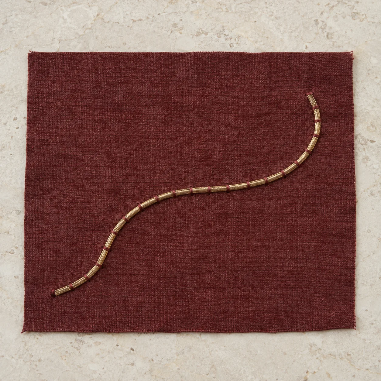 Gold-coloured metallic strand curved across maroon silk and held by thin maroon stitches
